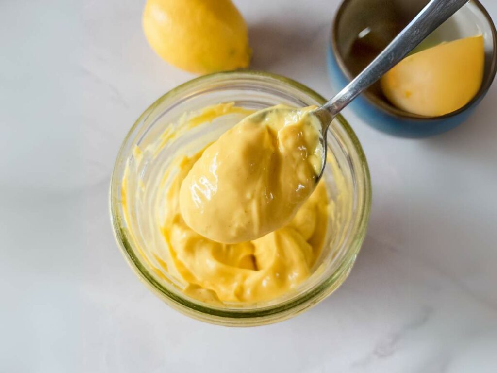 A spoon holding a dollop of yellow mayonnaise above a glass jar, with a lemon and a bowl in the background on a white surface.