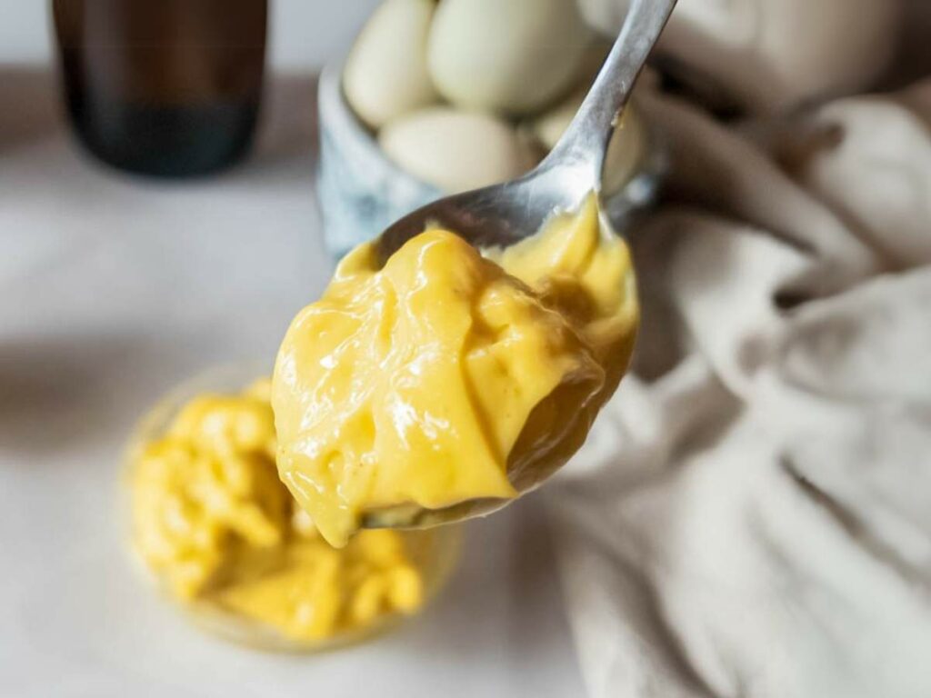 A close-up of a spoonful of creamy, yellow mayonnaise with a bowl of mayonnaise and whole eggs in the background.