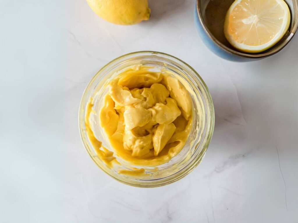 A glass jar filled with thick, yellow mayonnaise sits on a white surface next to a lemon and a bowl with a lemon slice.