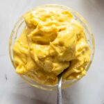 A glass bowl filled with thick, yellow mayonnaise with a spoon dipped into it, placed on a light-colored surface.