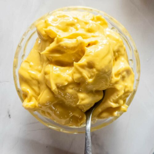 A glass bowl filled with thick, yellow mayonnaise with a spoon dipped into it, placed on a light-colored surface.