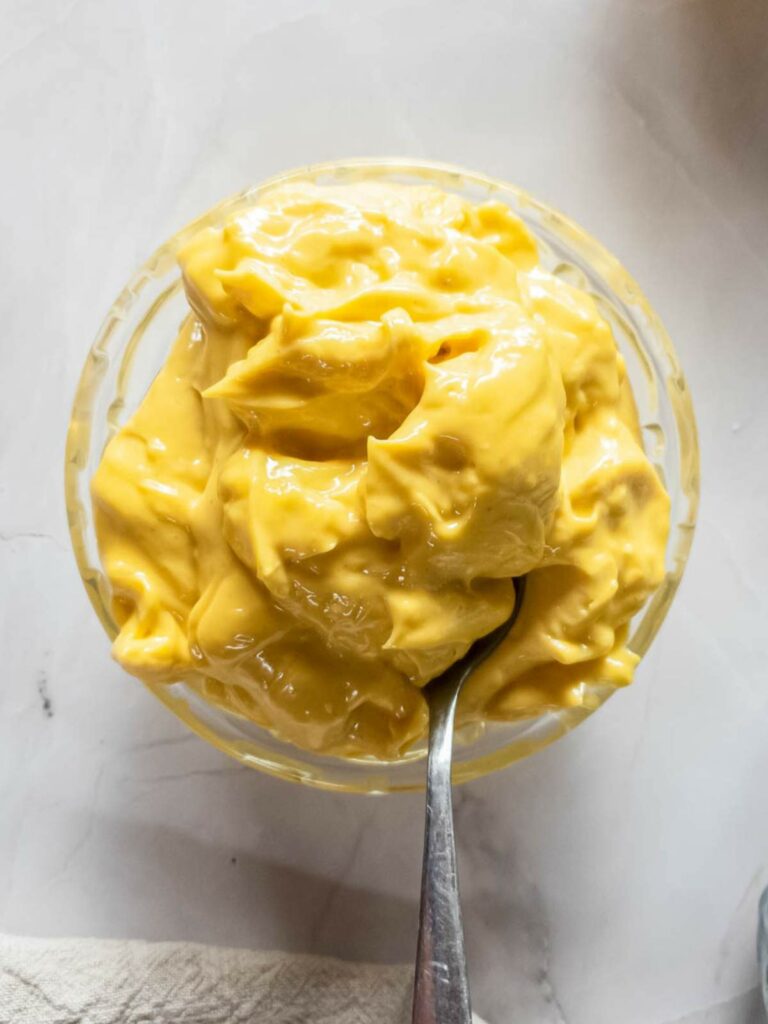 A glass bowl filled with thick, yellow mayonnaise with a spoon dipped into it, placed on a light-colored surface.
