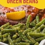 A close-up of cooked green beans topped with grated cheese on a white plate, with a piece of meat and a lemon wedge in the background.