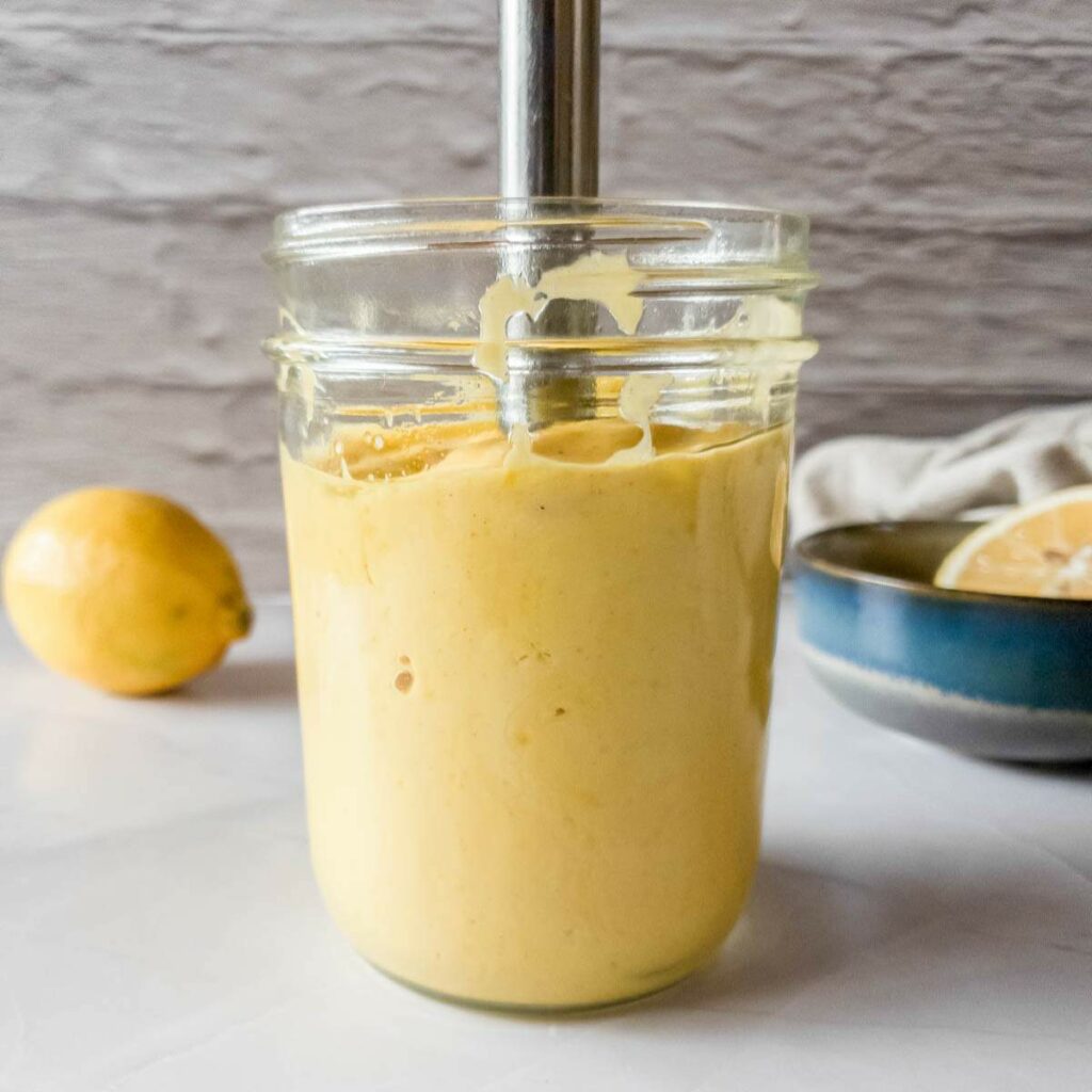 A glass jar filled with yellow mayonnaise being blended with an immersion blender.