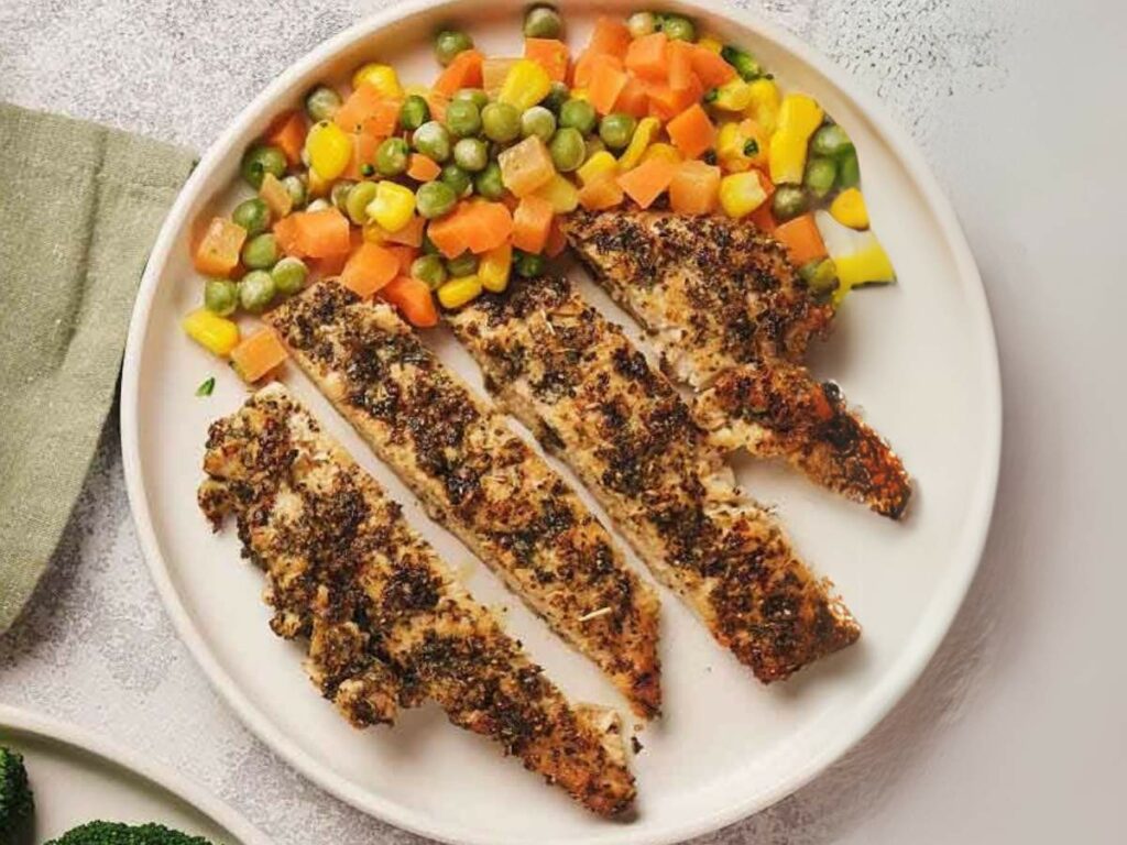 Grilled chicken strips with herbs are served on a plate alongside mixed vegetables including peas, corn, and diced carrots.