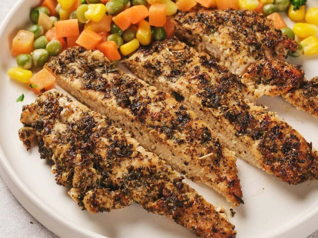 Sliced pan seared chicken breast with herbs, served with mixed vegetables including carrots, corn, and peas on a white plate.