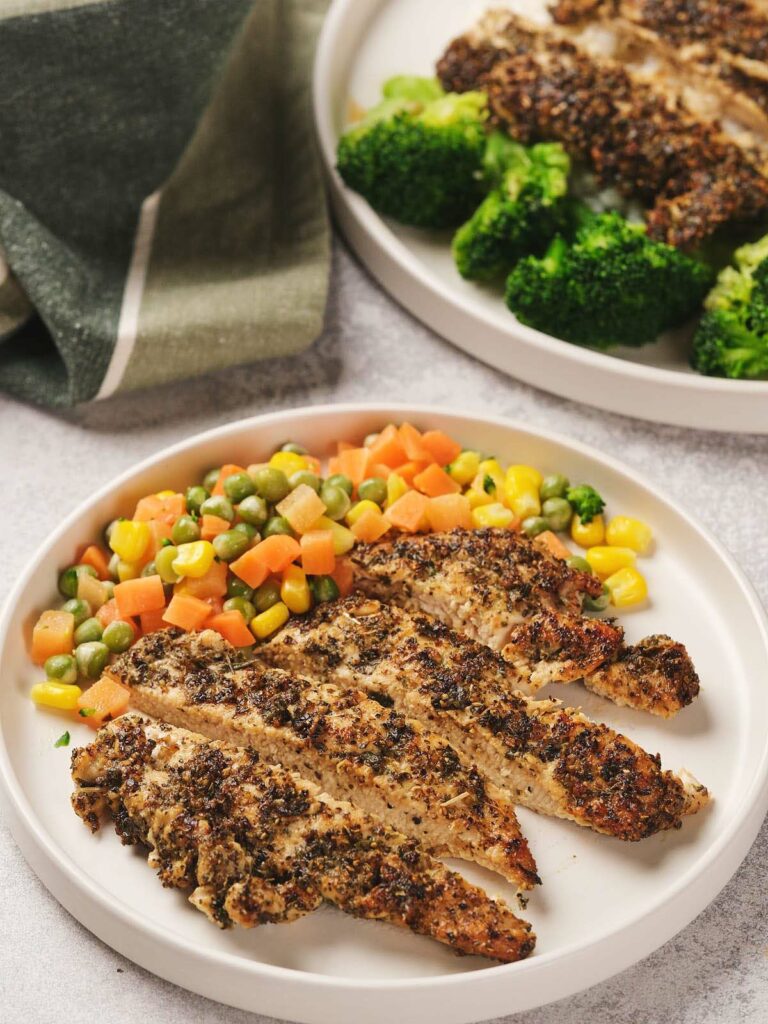 Pan seared chicken breast with mixed vegetables including carrots, peas, and corn on a plate, accompanied by broccoli in the background.