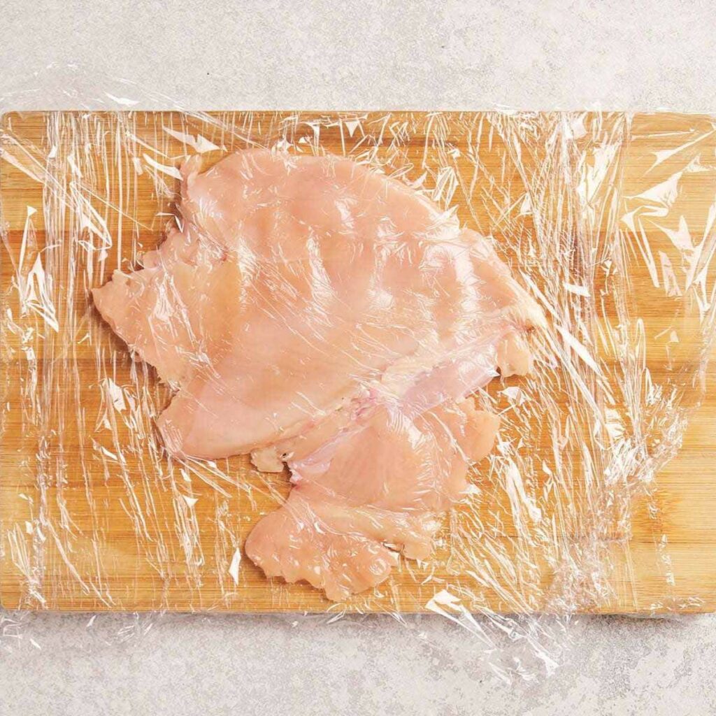 Raw chicken breast covered with plastic wrap on a wooden cutting board.