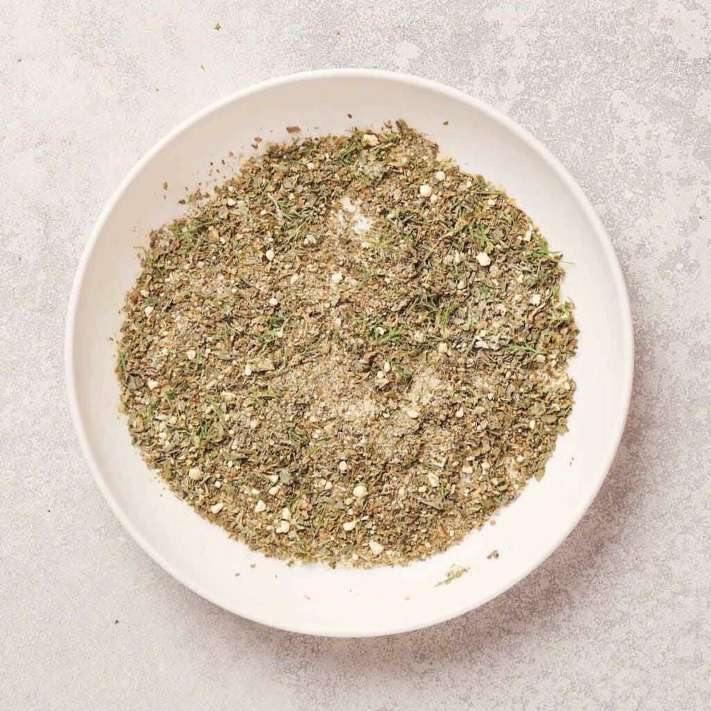 A white bowl filled with a mixed blend of dried herbs and seasonings on a light gray surface.