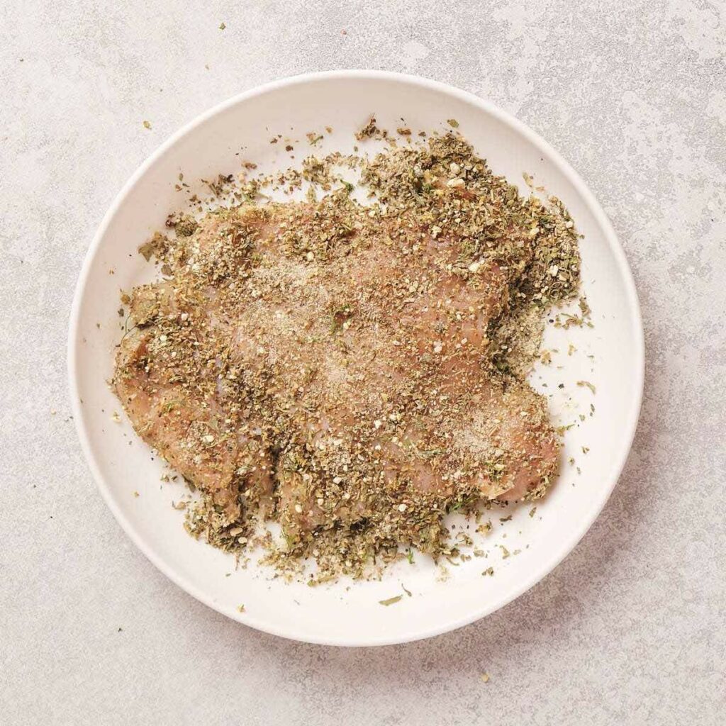 A piece of raw meat covered in a mixture of herbs and spices sits on a white plate against a light textured background.