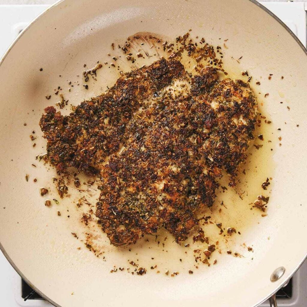 Seasoned chicken breast being pan-fried in a skillet with a herb and spice crust.