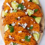 Quesabirria tacos filled with meat are arranged on a wooden board, garnished with chopped onions, cilantro, and lime wedges.