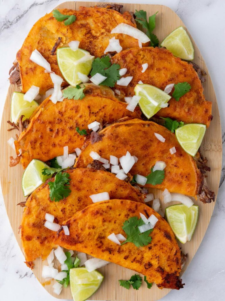 Quesabirria tacos filled with meat are arranged on a wooden board, garnished with chopped onions, cilantro, and lime wedges.