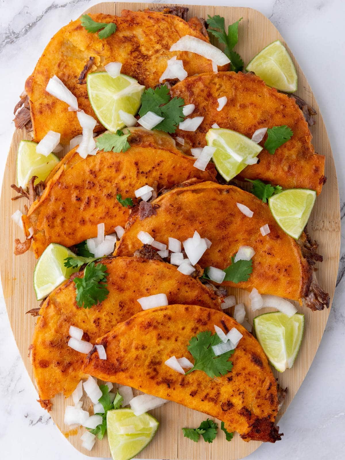 Quesabirria tacos filled with meat are arranged on a wooden board, garnished with chopped onions, cilantro, and lime wedges.