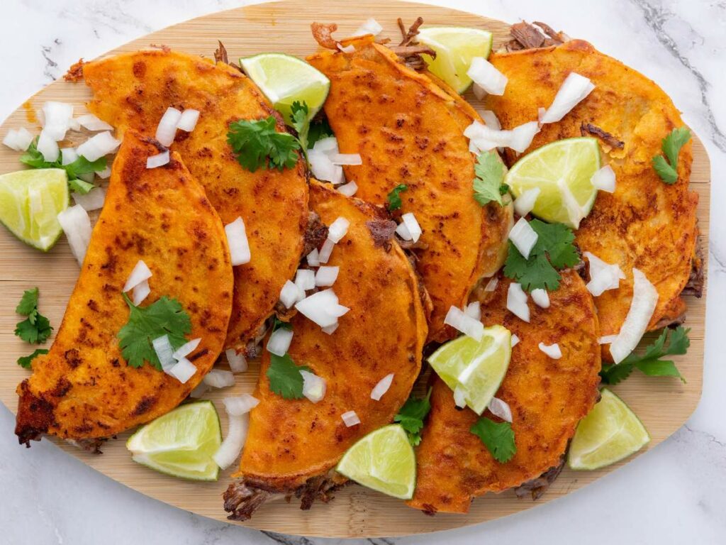 A wooden platter with several quesabirria tacoson a white surface.
