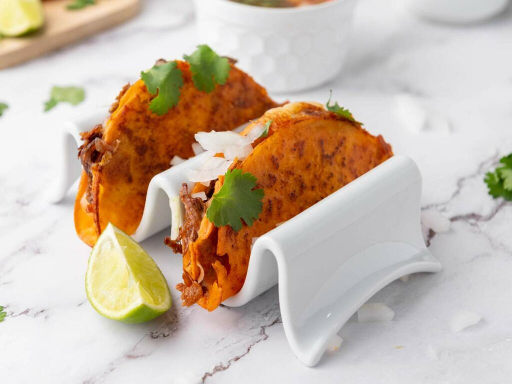 Quesabirria tacos in a white taco holder, garnished with chopped onions and cilantro, with a lime wedge.