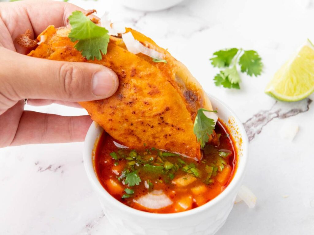 A hand dips a quesabirria taco filled with meat and cilantro into a bowl of birria consommé.