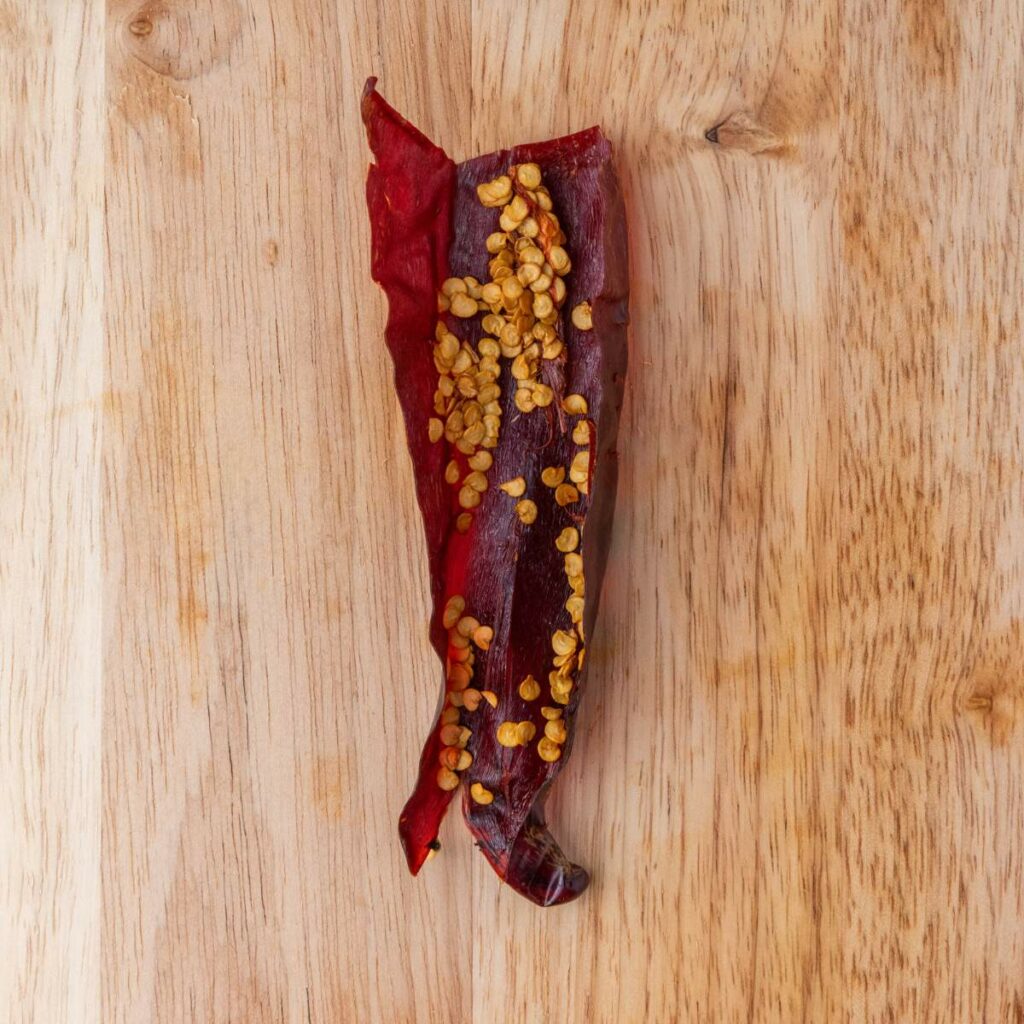 A dried red chili pepper with seeds is laying on a wooden surface.