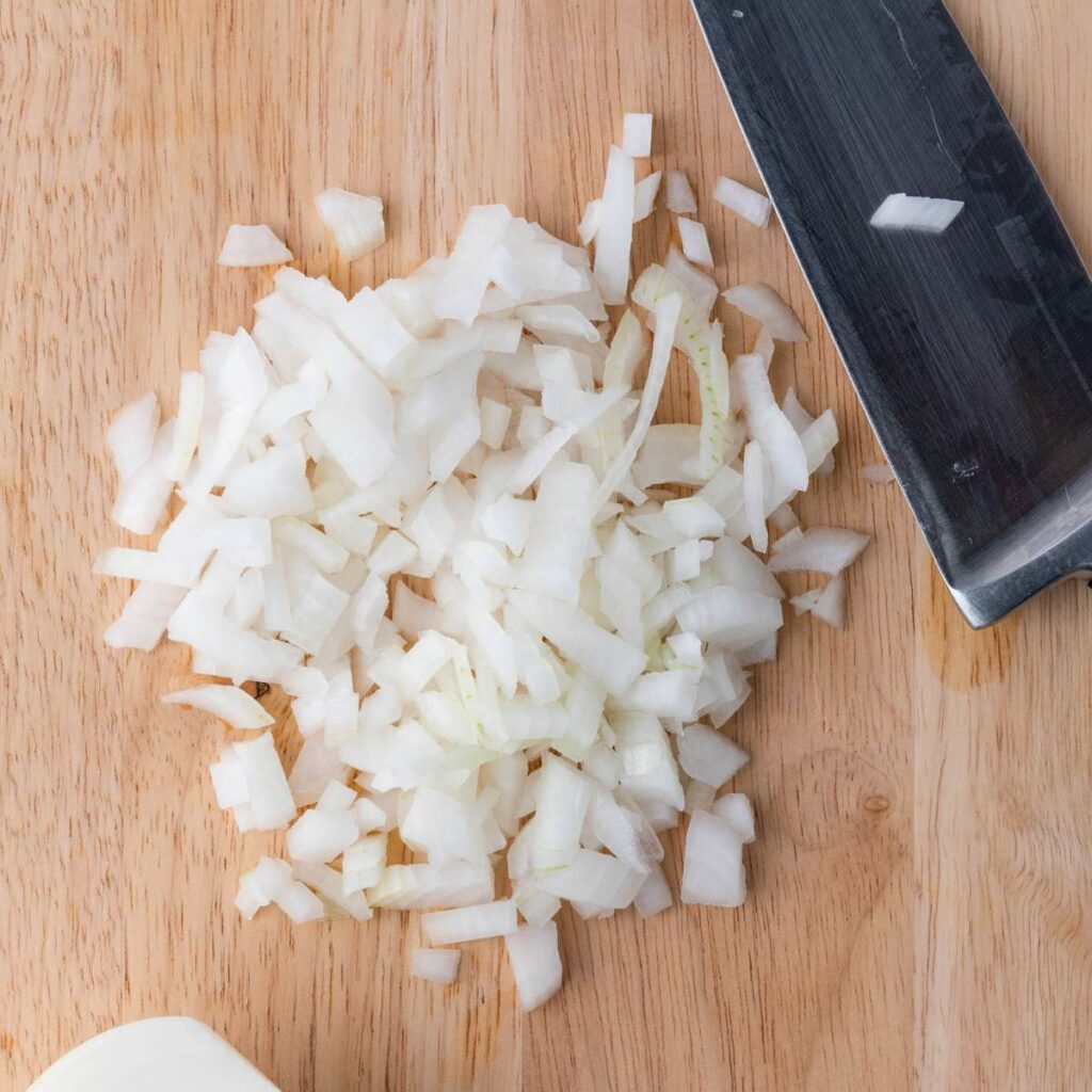 Finely chopped white onion on a wooden cutting board next to a kitchen knife.