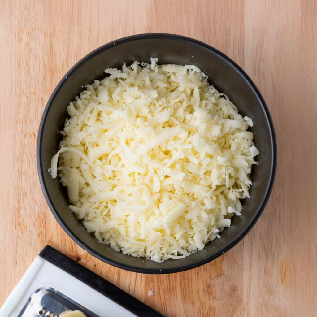 A black bowl filled with shredded cheese sits on a wooden surface next to a cheese grater.
