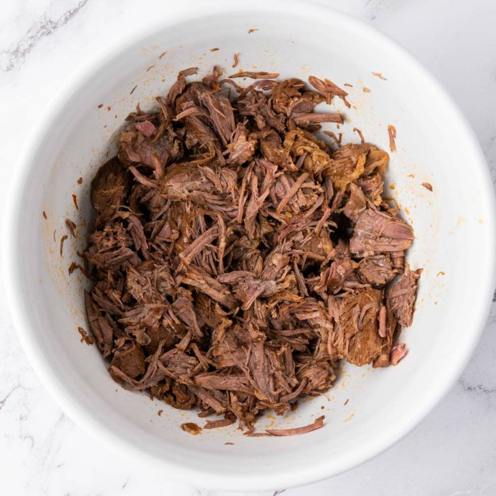 Shredded cooked beef in a white bowl on a marble countertop.
