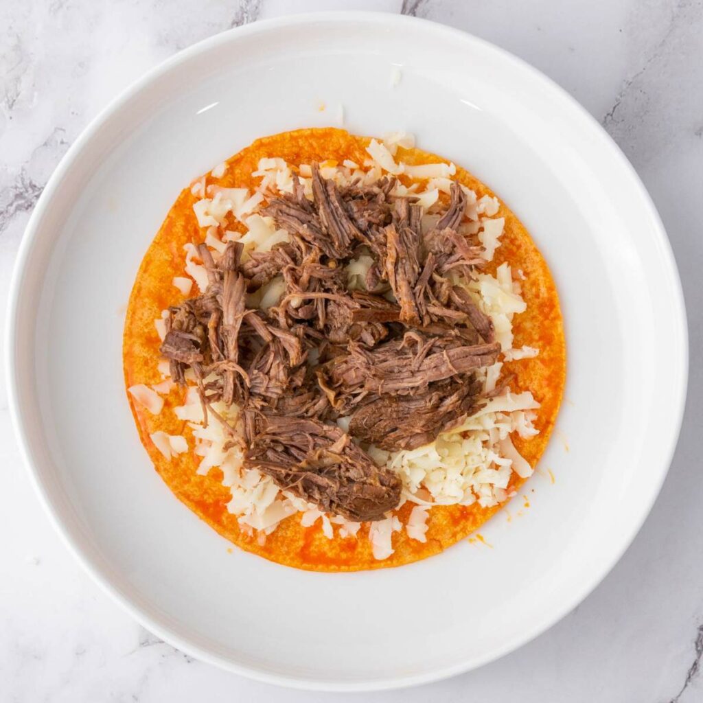 A white plate with a corn tortilla topped with shredded beef and melted cheese on a marble surface.