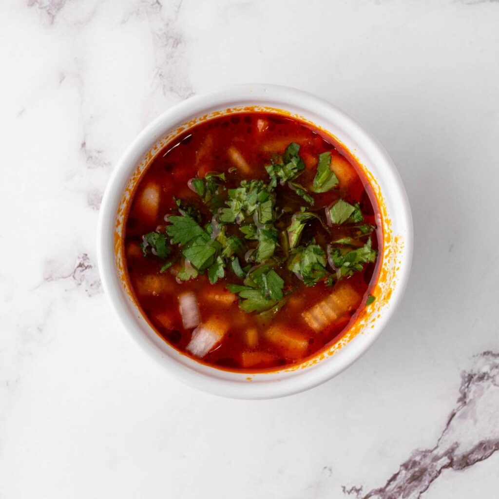 A bowl of red soup garnished with chopped onions and cilantro, placed on a white marble surface.