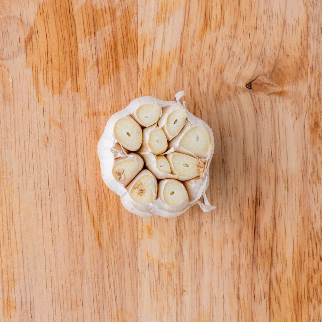 A head of garlic with the top cut off, exposing the cloves, is placed on a light wooden surface.
