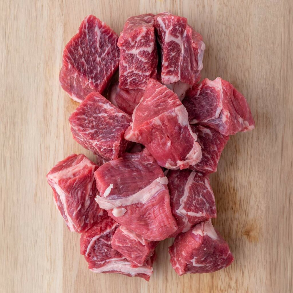 Chunks of raw beef with visible marbling arranged on a wooden cutting board.