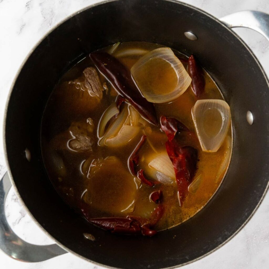 A black pot containing broth, halved onions, dried red chilies, and pieces of meat simmering on a stove.