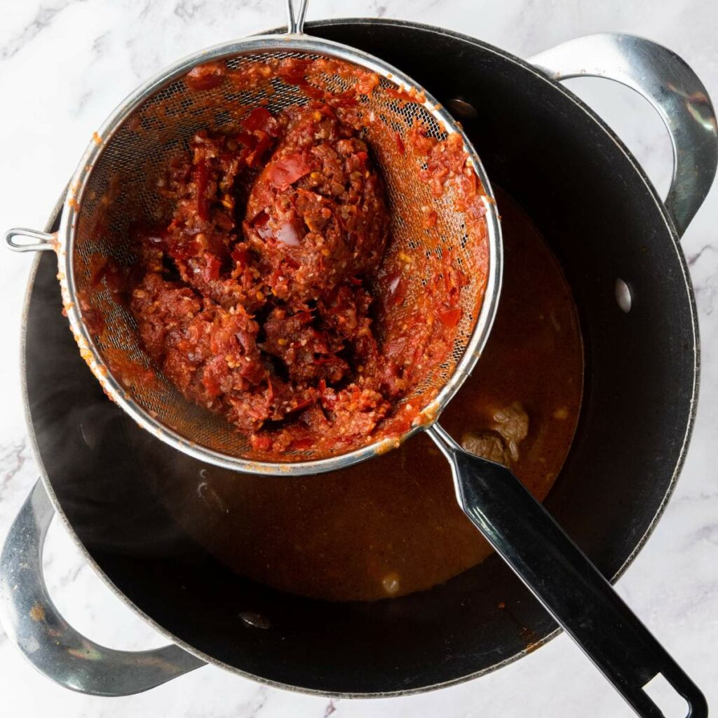 A metal strainer holding tomato paste is positioned over a black pot with broth on a marble countertop.