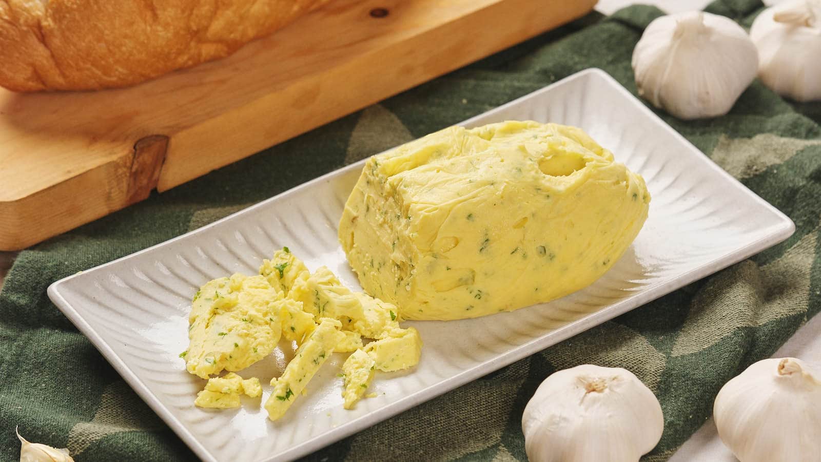A rectangular plate with roasted garlic butter on a cloth, surrounded by garlic bulbs, with a loaf of bread in the background.