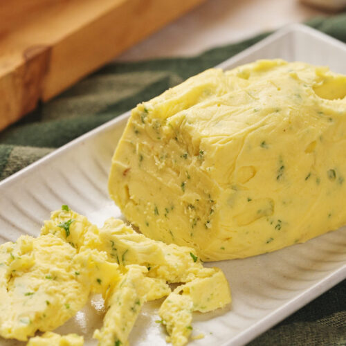 A rectangular white dish holds a partially sliced cylinder of roasted garlic butter.
