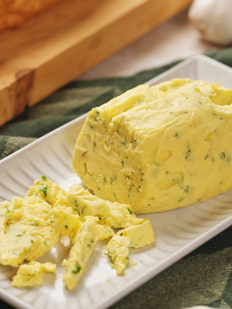 A rectangular white dish holds a partially sliced cylinder of roasted garlic butter.