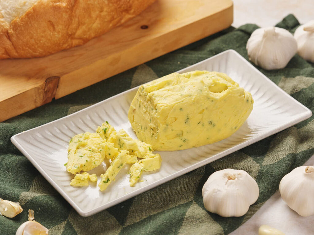 Roasted garlic butter on a rectangular white plate is placed on a green cloth, surrounded by garlic bulbs.