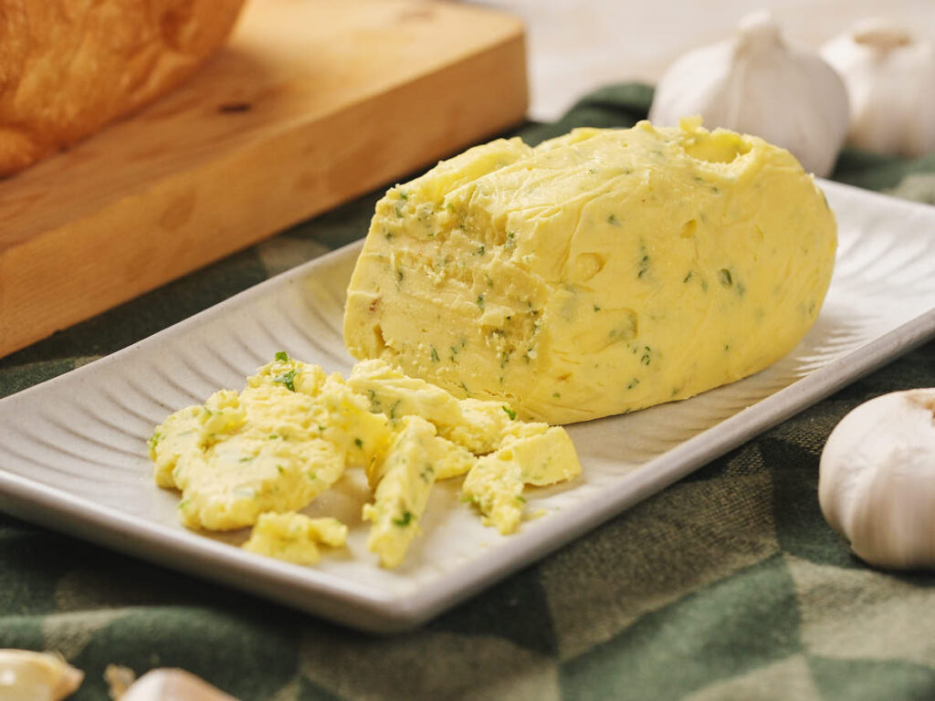 Roasted garlic butter sits on a ceramic plate, surrounded by garlic cloves on a green checkered cloth.