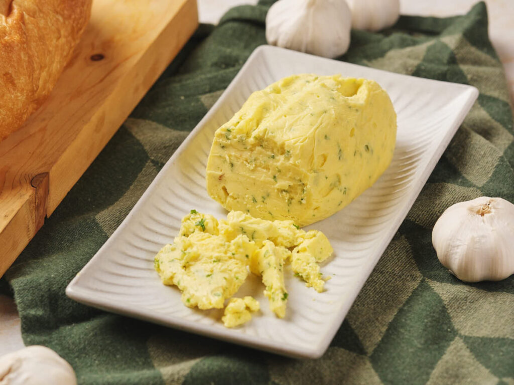 A rectangular dish with roasted garlic butter, some of it crumbled. Surrounded by garlic cloves on a green cloth.