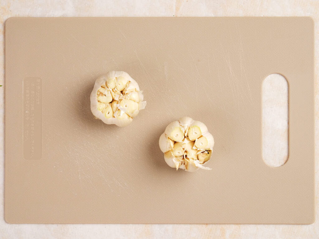 Two halved garlic bulbs on a beige cutting board with a handle, placed on a light surface.