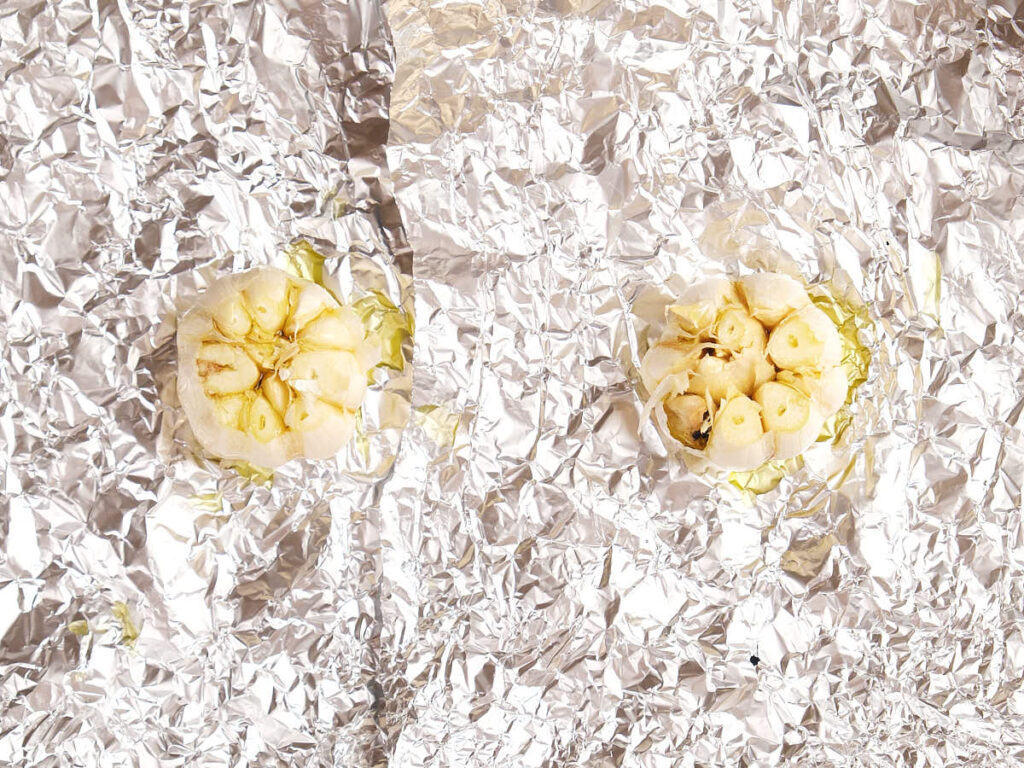 Two garlic bulbs with tops cut off placed on crinkled aluminum foil, ready for roasting.
