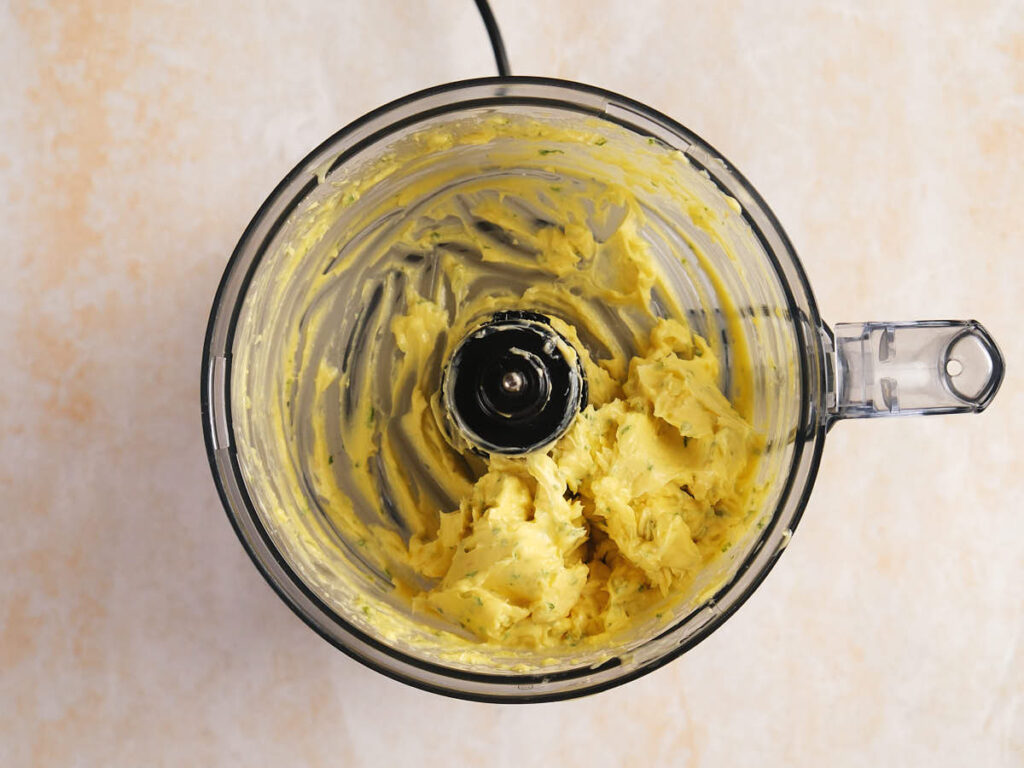 Food processor bowl with blended yellow butter mixture at the bottom, showing streaks on the sides.