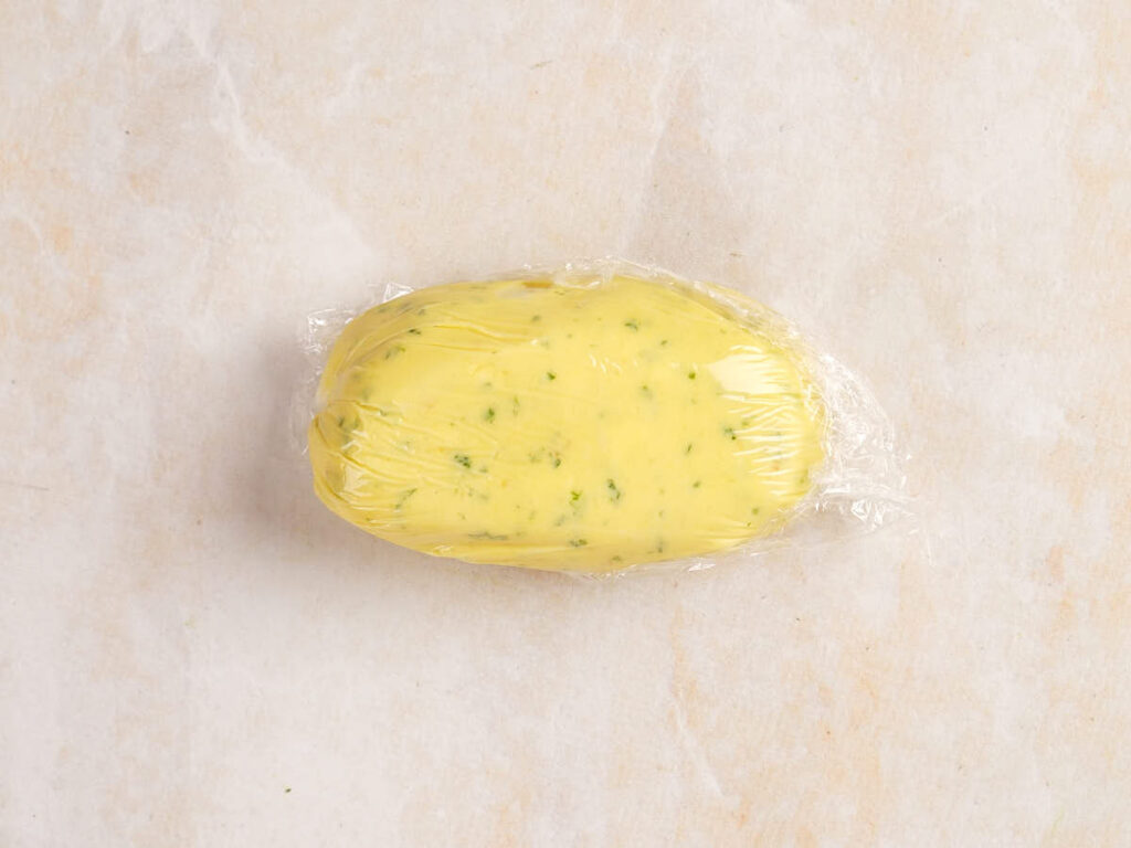 A piece of yellow herbal butter wrapped in plastic wrap on a light marble surface.