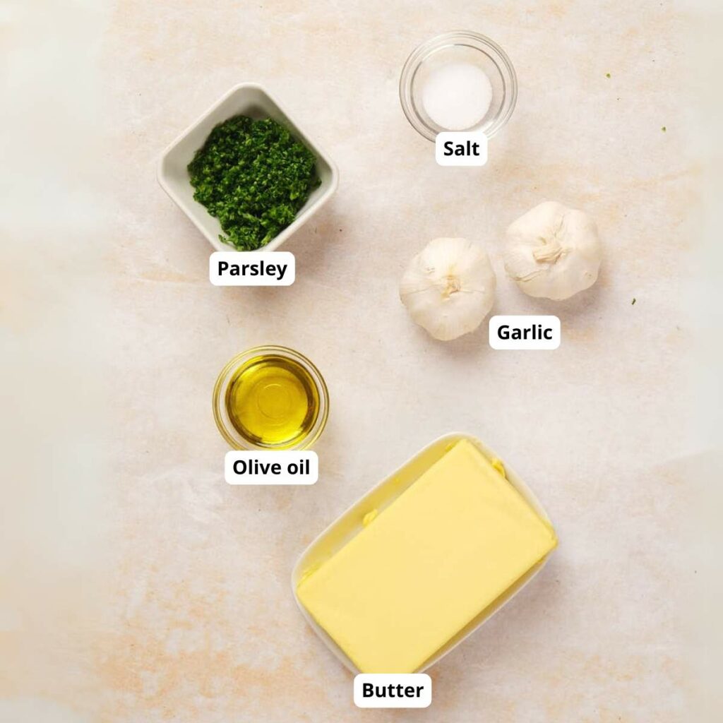 Butter, olive oil, parsley, salt, and two garlic bulbs are arranged on a light surface, each labeled with text.