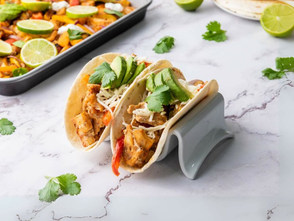 Two chicken fajitas with avocado, cheese, and cilantro in a taco holder on a marble surface.