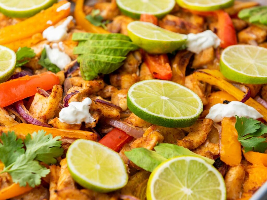 Sheet pan chicken fajitas topped with lime slices, avocado, cilantro, and dollops of sour cream.