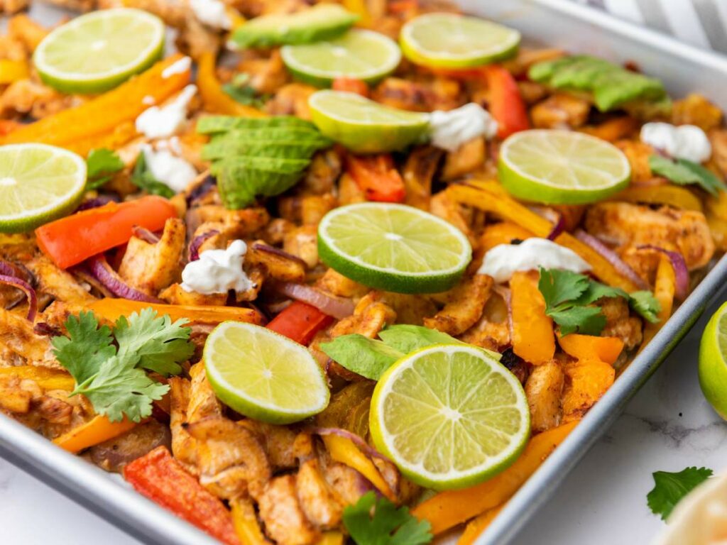 Sheet pan with cooked chicken fajitas, sliced bell peppers, red onions, lime slices, cilantro, avocado, and dollops of sour cream.