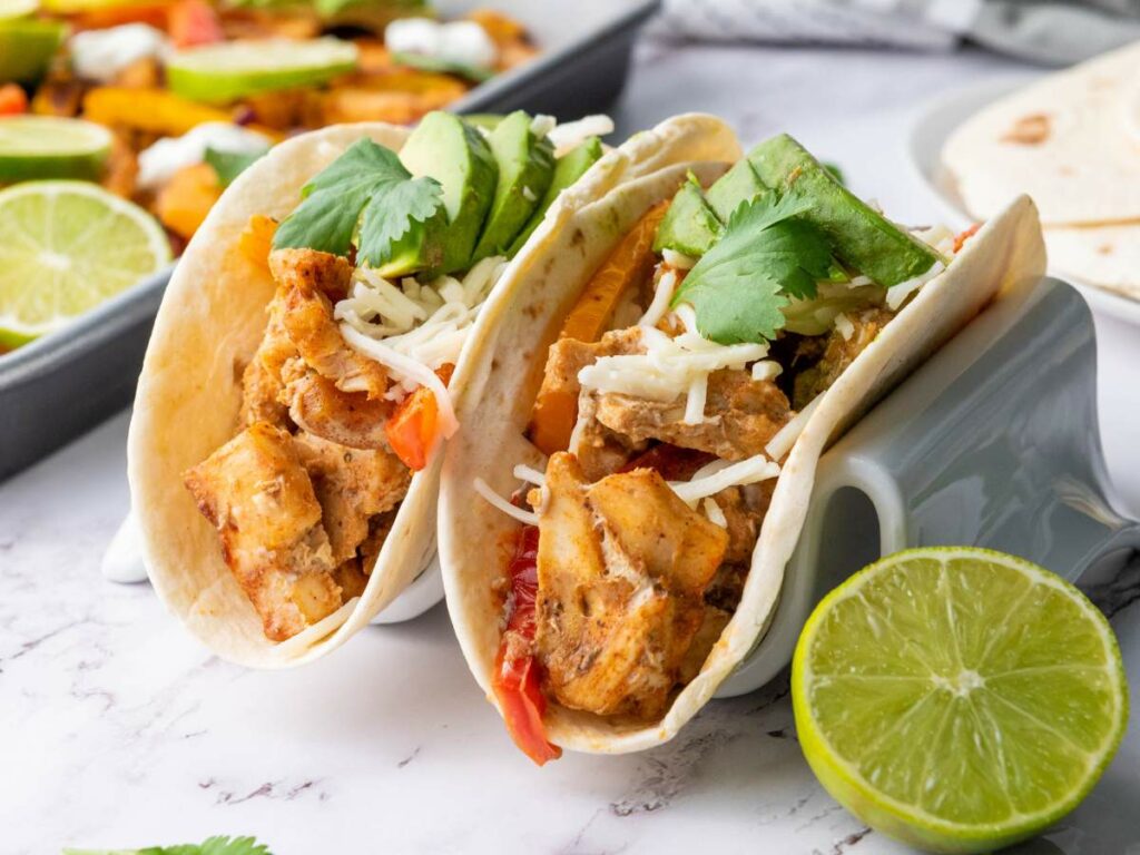 Two chicken fajitas in tortillas topped with shredded cheese, avocado slices, and cilantro, with a halved lime beside them on a white surface.
