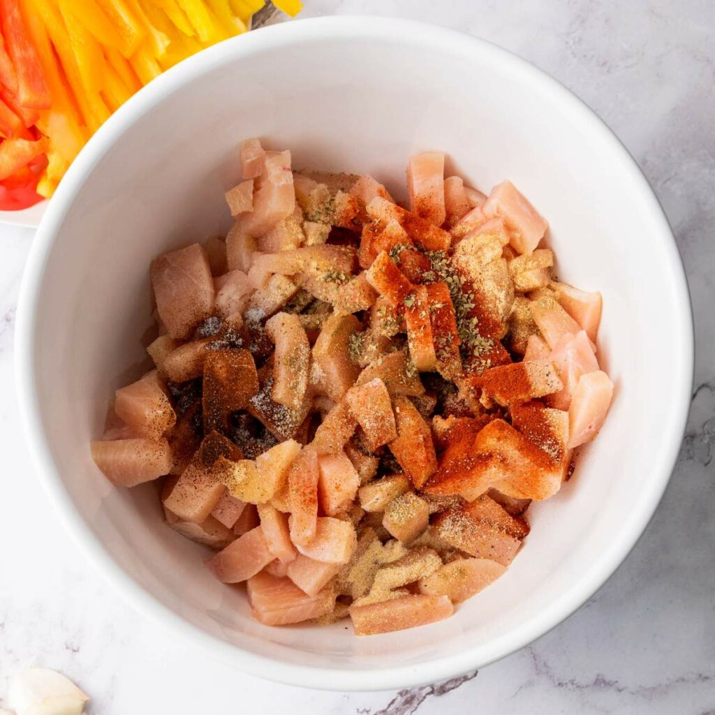 A white bowl containing diced raw chicken seasoned with various spices, with chopped yellow and red bell peppers visible in the background.
