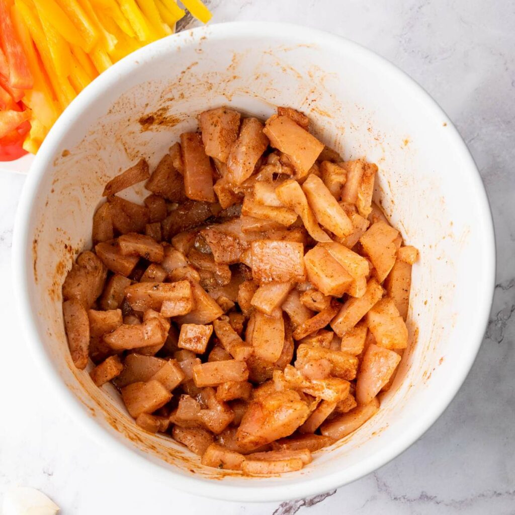 A white bowl filled with seasoned, uncooked chicken pieces on a marble surface.