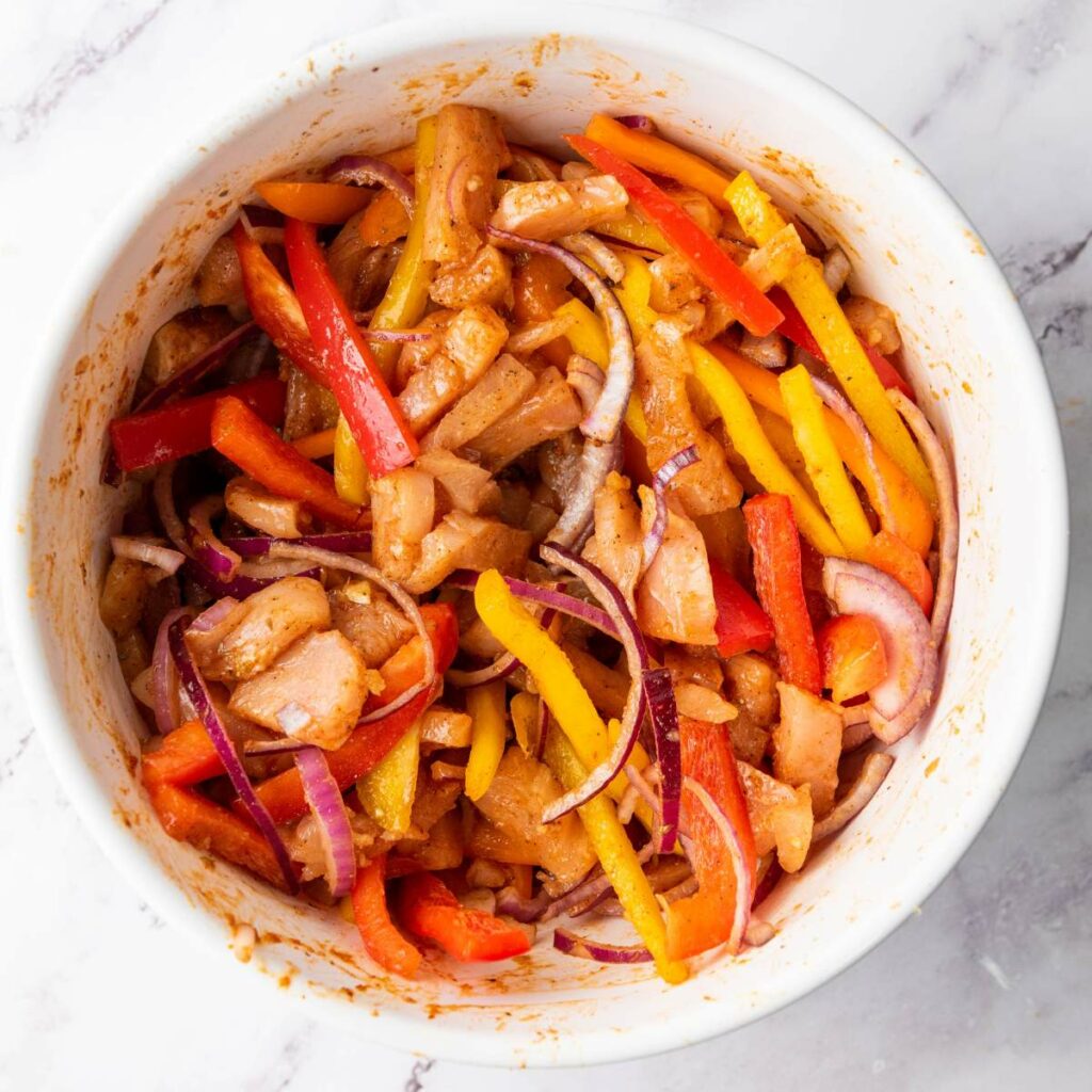 Bowl filled with sliced raw chicken, red onions, red and yellow bell peppers, and seasoning, ready to be cooked.