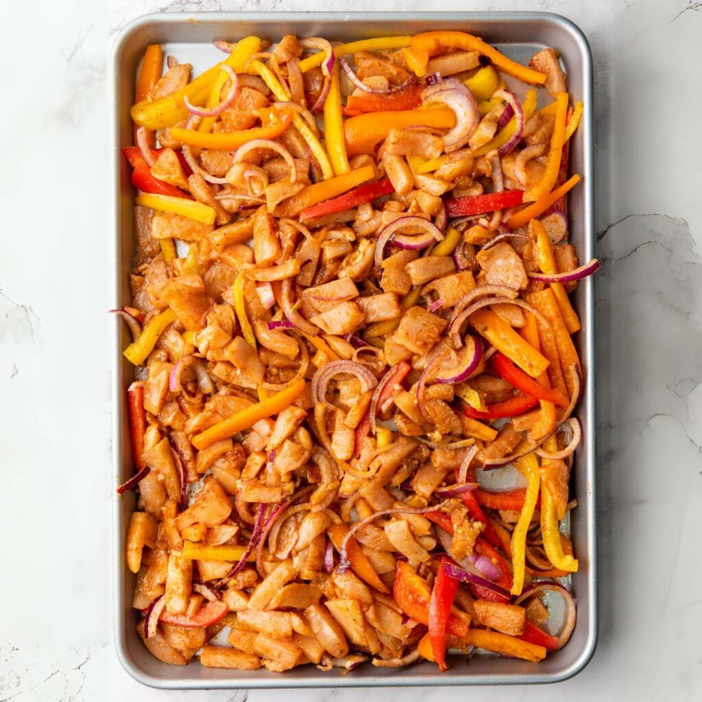 Sheet pan filled with sliced chicken, red onions, and assorted bell peppers, all coated in seasoning, ready to be cooked.